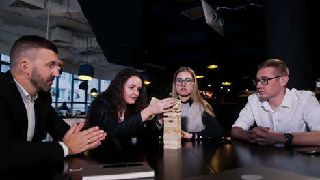 four people sit at the table with jenga block pile on it. dark-haired girl pulls the block very carefully while others watch her.