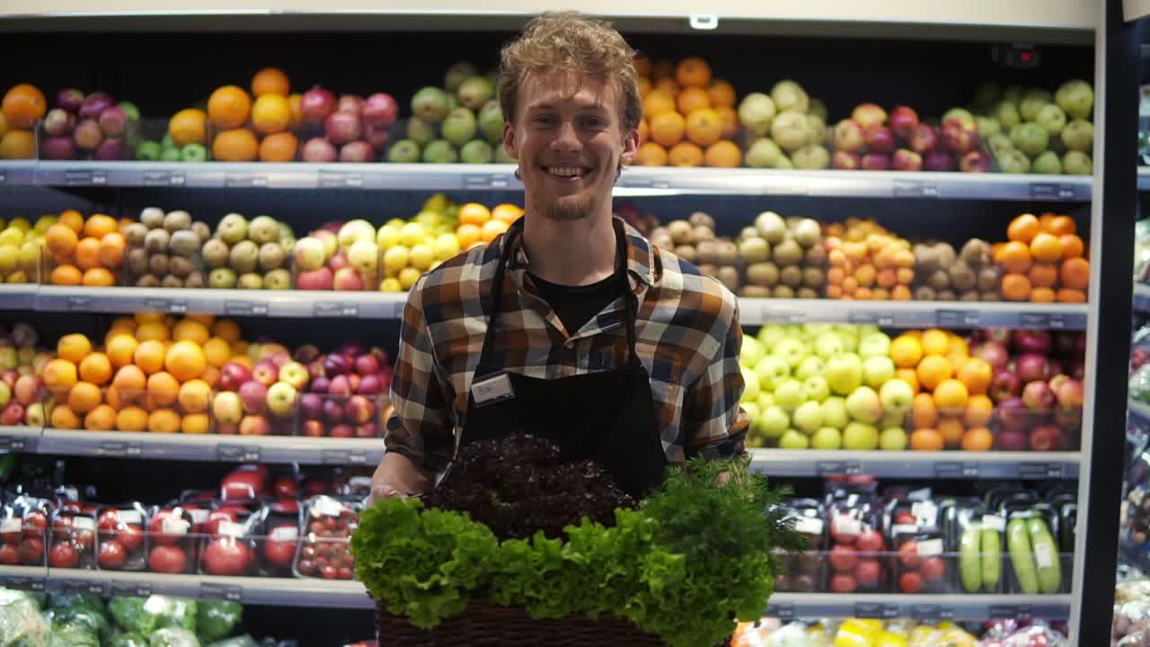 Portrait shot of the young handsome Caucasian shop worker in the apron standing in front the camera and smiling joyfully while holding box coloured greens at the grossery supermarket. Fruits and vegetables panel background