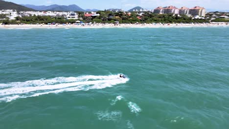 drone aerial scene of person riding jet ski doing water sports on beach near sand with blue sea in jurere internacional