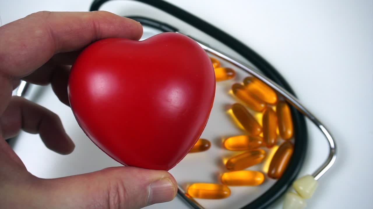 Persona tiene un cuore rosso sopra pillole di omega 3 e uno stetoscopio su uno sfondo bianco