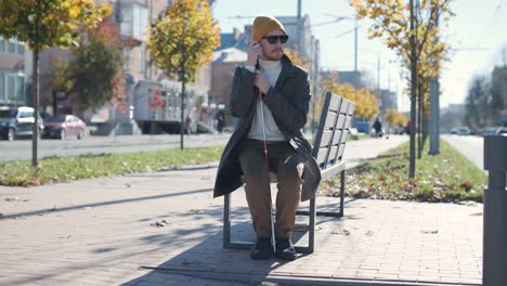 young blind man with smartphone sitting on bench in park in city, calling