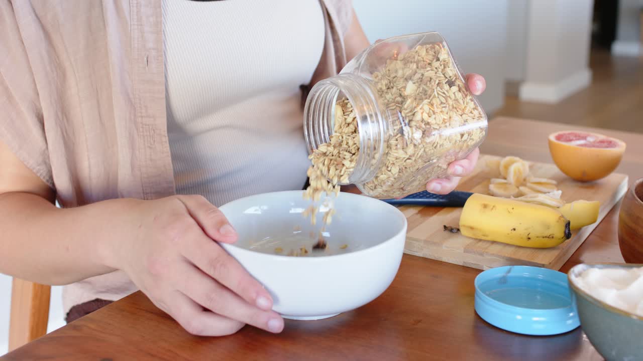 Donna afroamericana toglie un coperchio blu versando muesli da un barattolo in una ciotola al tavolo mentre prepara la colazione.