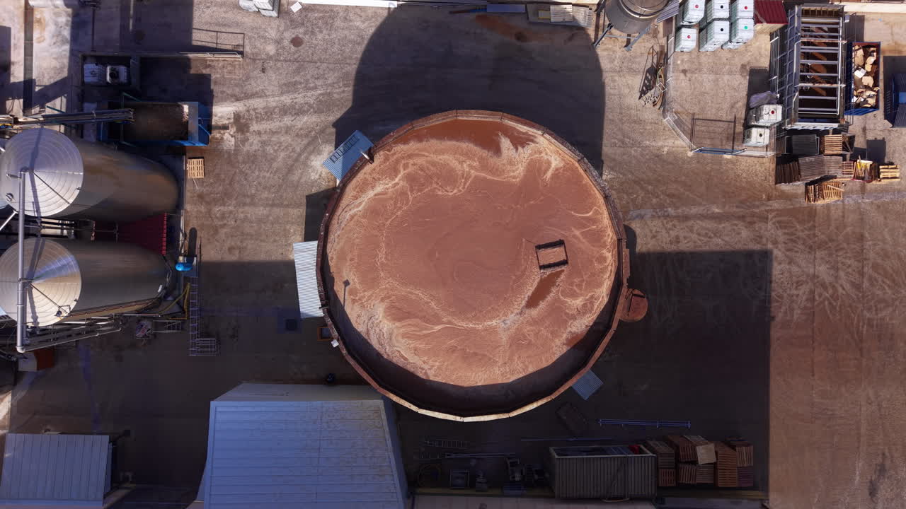 Vista de cima de um grande tanque clarificador industrial circular preenchido com água turva marrom-avermelhada em uma instalação de processamento.