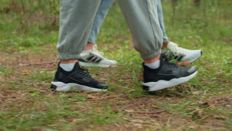 close up leg view of two individuals wearing sneakers walking on forest trail with visible green grass, pine needles, and soft natural light creating peaceful atmosphere during