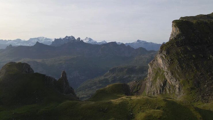 A drone flies towards Gross Achslenstock, capturing a breathtaking aerial view of rugged mountain peaks and lush green valleys under a clear sky, showcasing the majestic beauty of the landscape