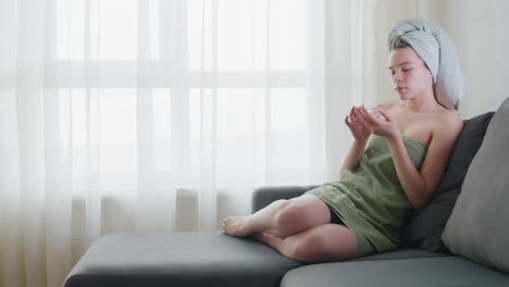 young lady wrapped in green towel sits with legs crossed on couch, gently smelling liquid skincare product and applying it beside nose in bright room with soft natural light