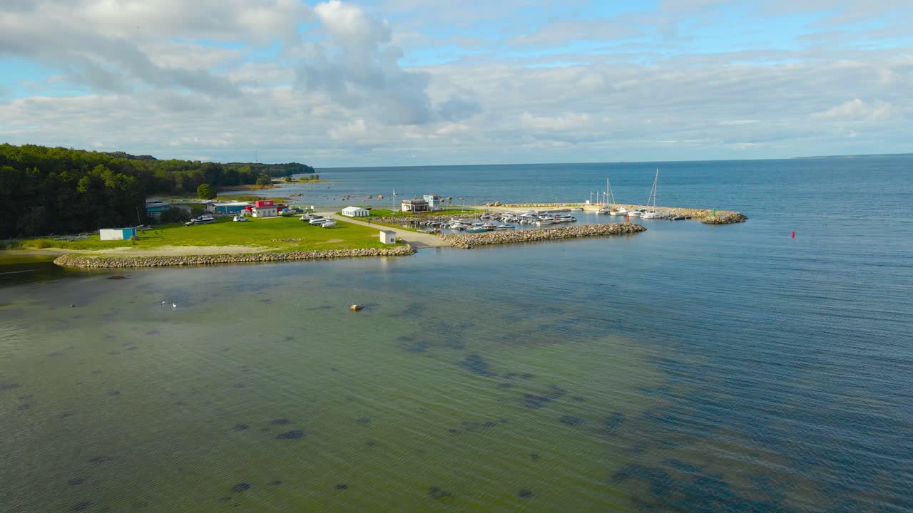 Aerial drone footage of a luxury and modern Tilgu yach club harbor where boats and sailboats are visible docked on the rocky pier during a sunny summer day with shallow Baltic sea water around it