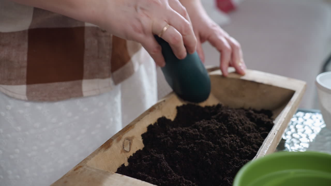 Lower view showing close up of hands with gold ring mixing rich loam soil using dark green plastic scoop in wooden planter box placed on glass table next to empty pot under soft natural light