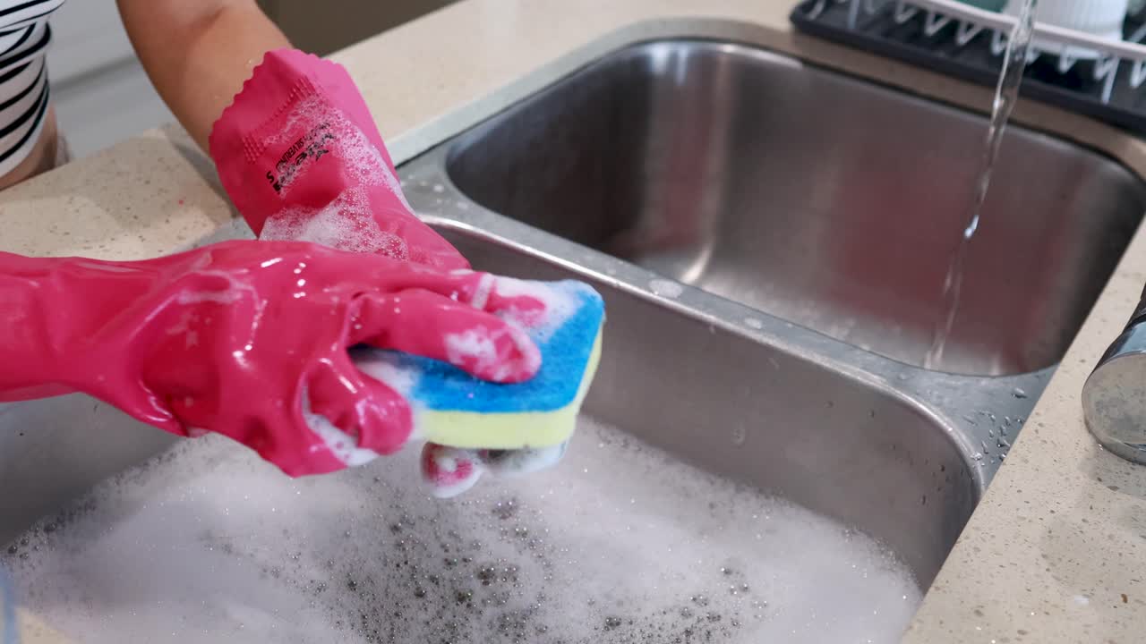 A person wearing pink gloves cleans a spoon using a sponge in a kitchen sink filled with soapy water