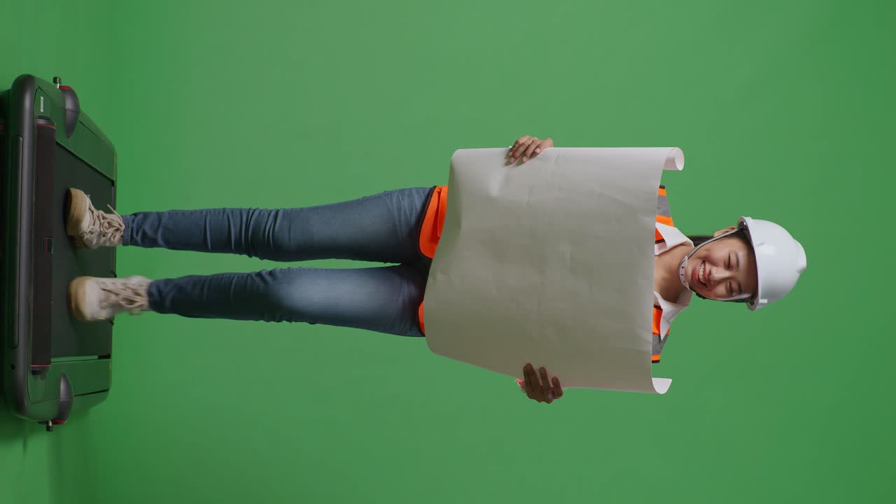 cuerpo lleno de ingeniera femenina asiática con casco de seguridad sosteniendo el plano en sus manos y mirando a su alrededor mientras camina en el estudio de fondo de la pantalla verde