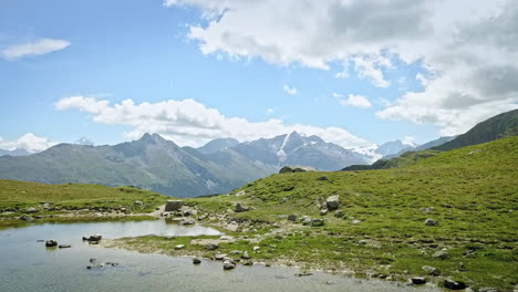 vuelo aéreo sobre un estanque pacífico con agua clara en el prado suizo revela la majestuosa ladera de la montaña de los alpes suizos mientras grandes nubes se mueven por el cielo