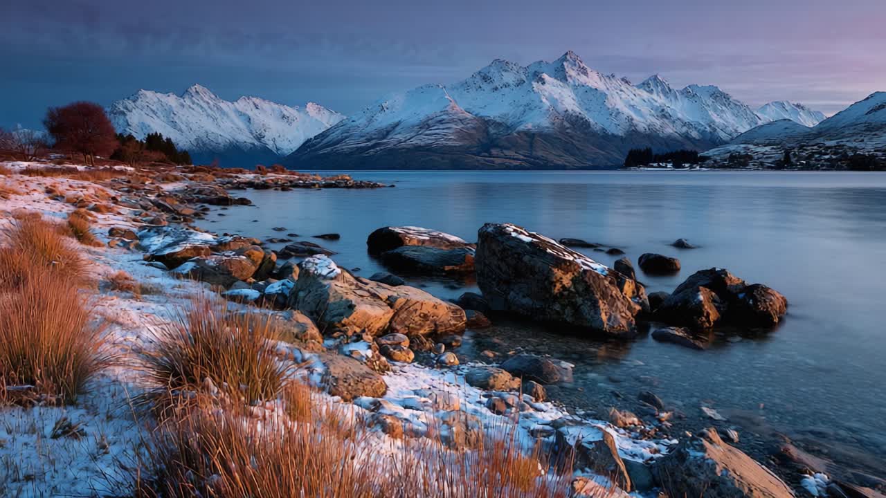 Un tramonto sereno su montagne innevate che si riflettono in un lago calmo, circondato da sassi ed erba, che evoca un senso di tranquillità e bellezza naturale
