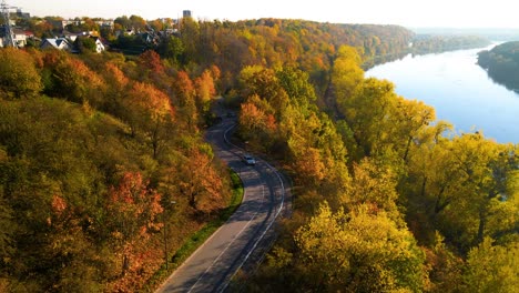 droneshot of a car driving on winding road adjacent to railway alongside nemunas river in kaunas, lithuania, zoom in shot