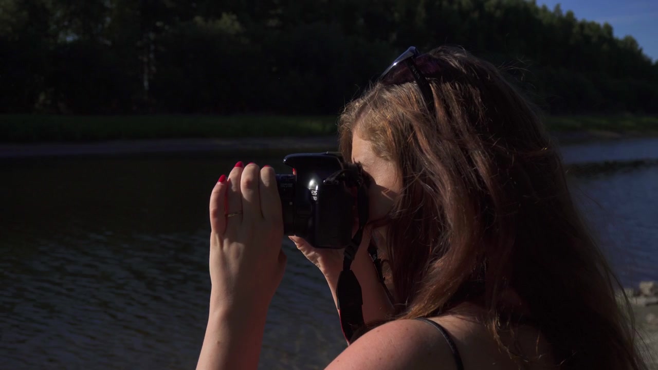 Young girl is taking photos with her dslr camera at nature park with river clear blue sky and forest in the background.
