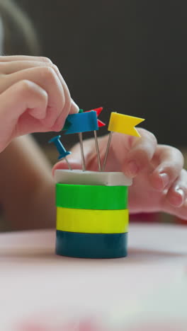 little girl decors small tower with tag pins and shows hearts by fingers at desk in classroom closeup. student crafts of stationery in playroom