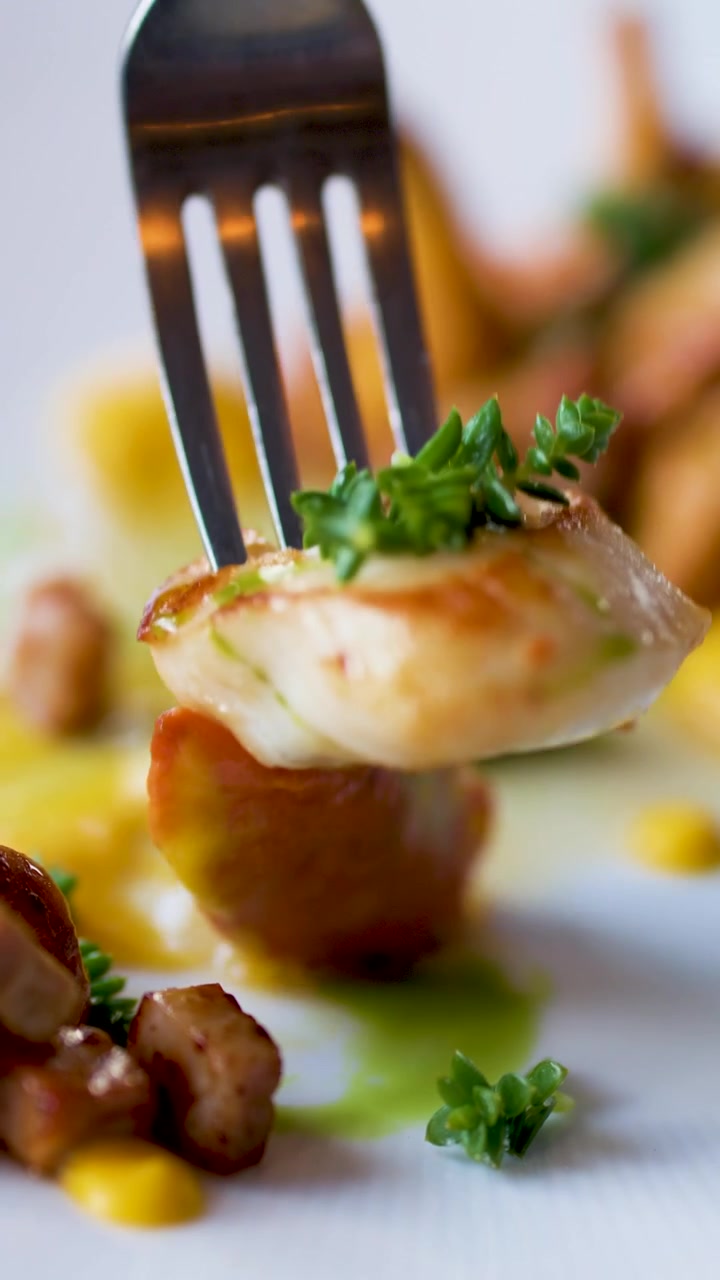 Una sequenza ravvicinata mostra una forchetta che trafigge e solleva una capasanta scottata guarnita con erbe aromatiche da un elegante piatto di pesce gourmet sotto una luce brillante e naturale.