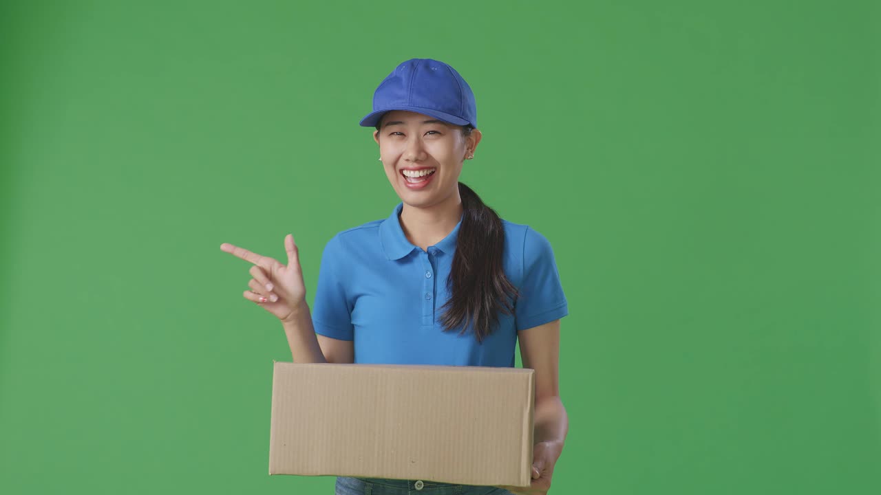 Asian Female Courier In Blue Uniform Smiling And Pointing To The Side While Delivering A Carton On Green Screen Background In The Studio
