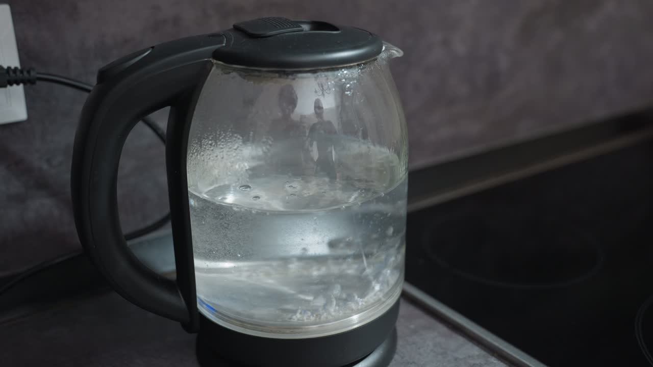 Elektrischer Glaswasserkocher kocht auf der Arbeitsfläche, blubberndes Wasser mit aufsteigendem Dampf und blauem LED-Sockel, aktive Erwärmung mit lebhafter Blasenbewegung im Inneren des klaren Glases, Frühstücksszene für Barista oder Büro