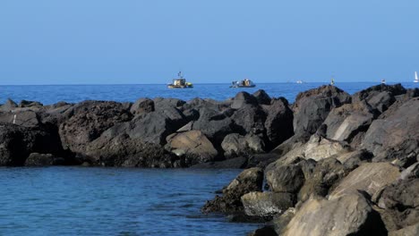 Stone-pier-at-the-beach-of-Playa-De-Las-Americas-,-sunny-summer-day,-calm-Atlantic-ocean-in-the-background,-distant-fishing-boats,-medium-handheld-shot