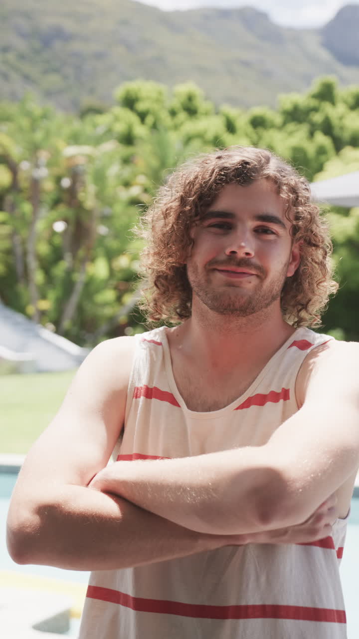 Vertical video: Smiling man with curly hair standing by pool with arms crossed
