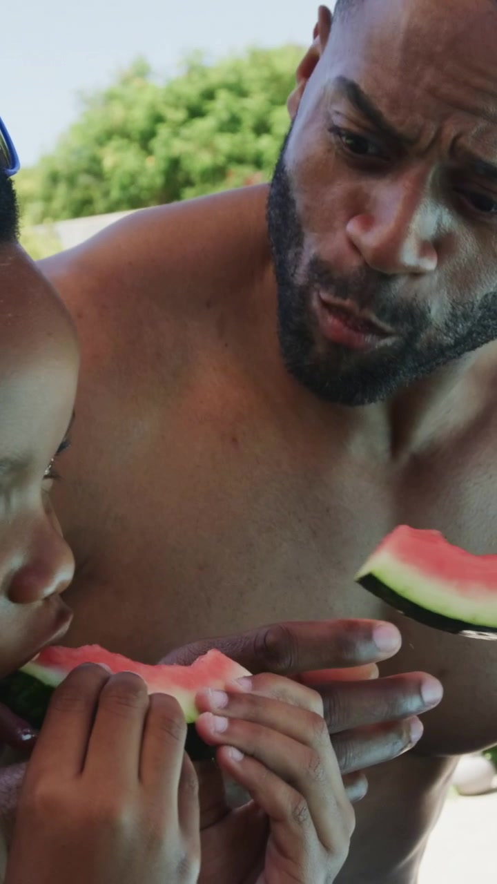 Vertical video: Picking watermelon, shirtless dad snacking poolside while child with blue goggles