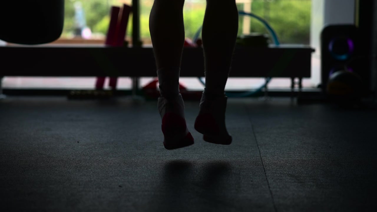 Low angle view shows lower legs and feet in athletic shoes and socks lifting slightly above dark floor inside fitness room, with strong backlight, soft blur, quiet workout atmosphere, and steady