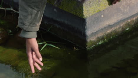 Cerrar-Una-Mujer-Joven-Salpicando-La-Mano-En-El-Agua-Tocando-Juguetonamente-Un-Estanque-Fresco-En-Un-Parque-Natural-Al-Aire-Libre