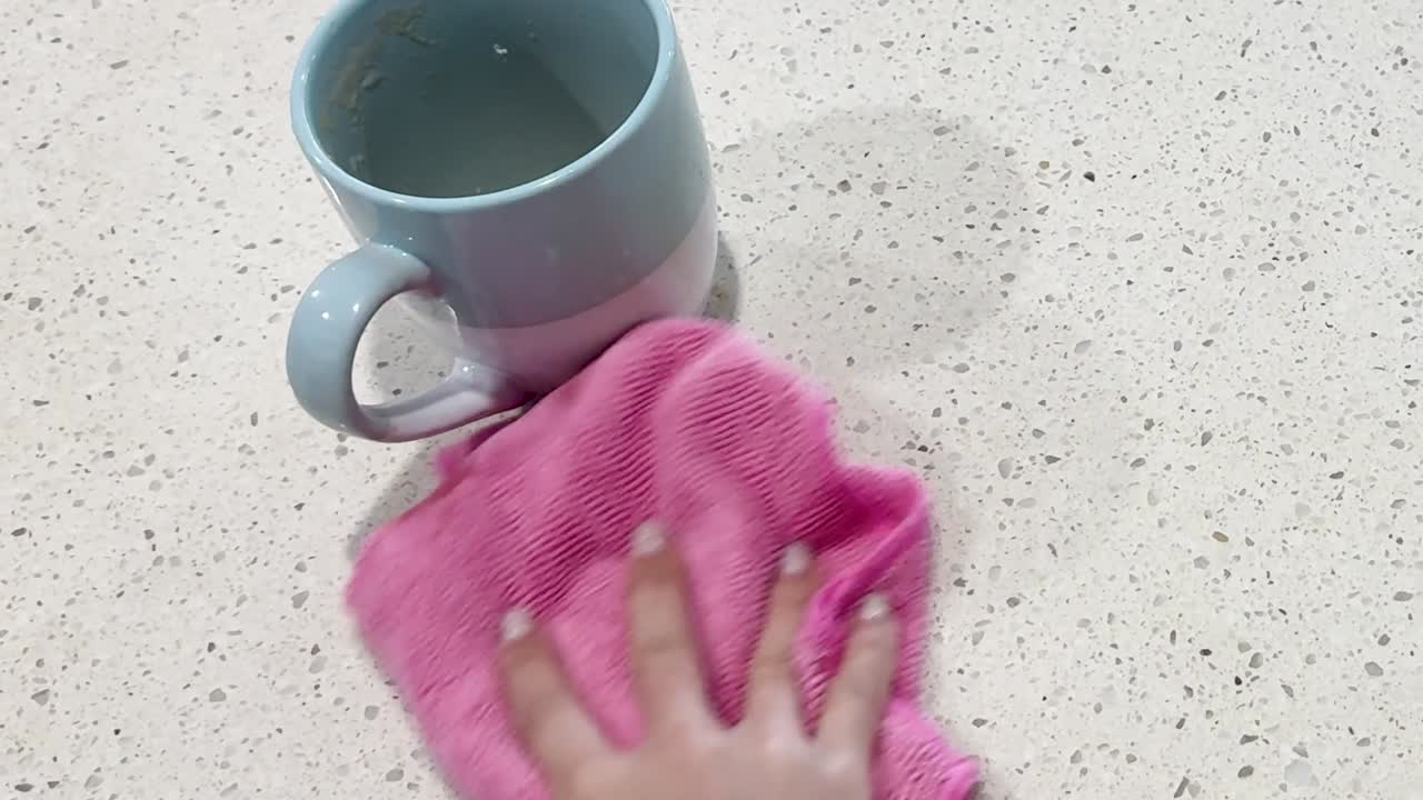 A hand uses a pink microfiber cloth to wipe a liquid spill on a countertop