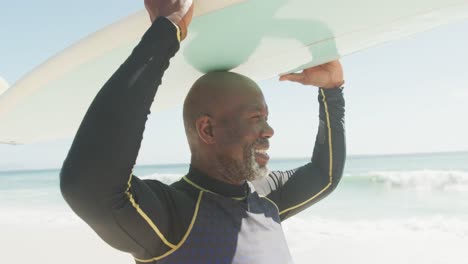 portrait of smiling senior african american man walking with surfboard on sunny beach