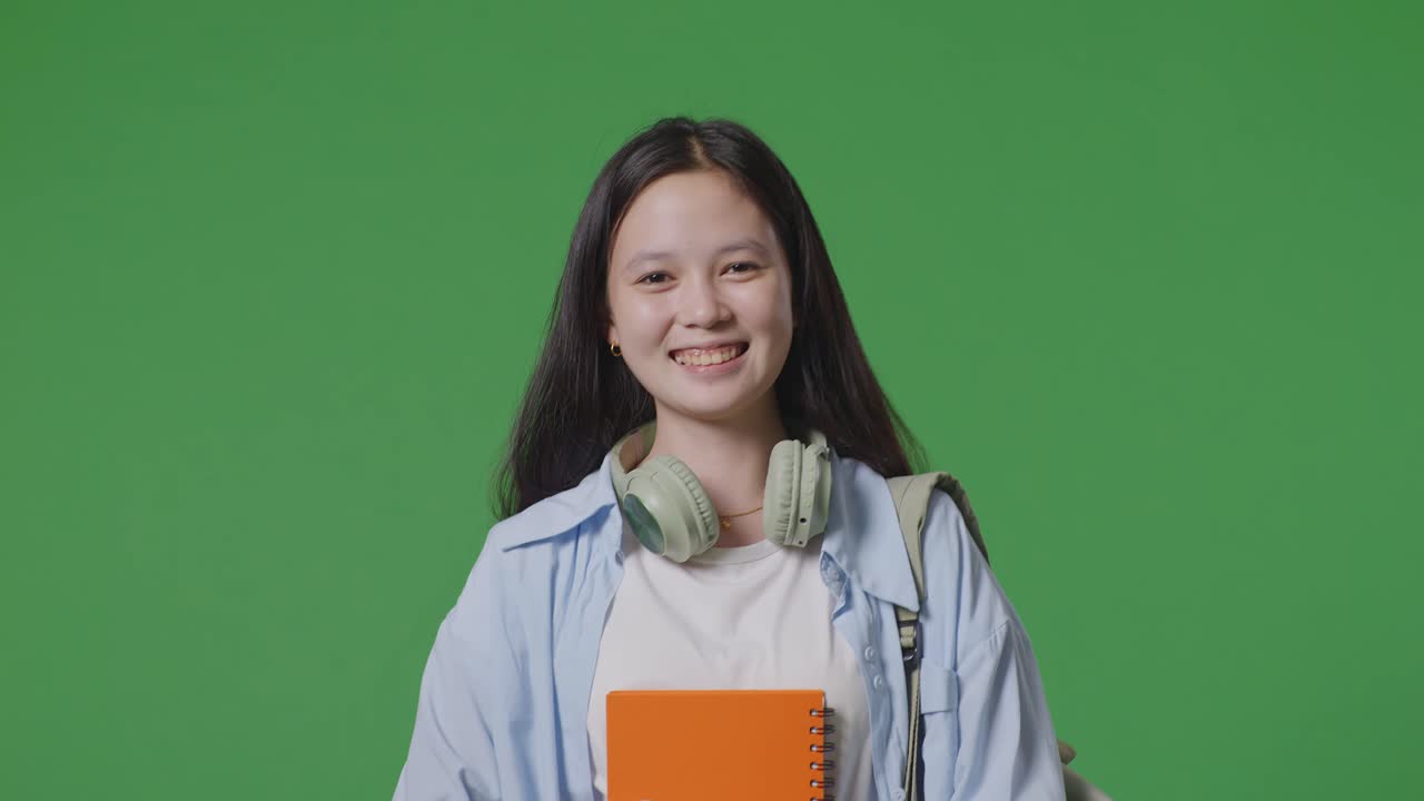 アジアのティーンエイジャーの女の子の学生がバックパックを背負っていくつかの本を握り緑のスクリーン背景のスタジオで歩いている間に微笑んでいます