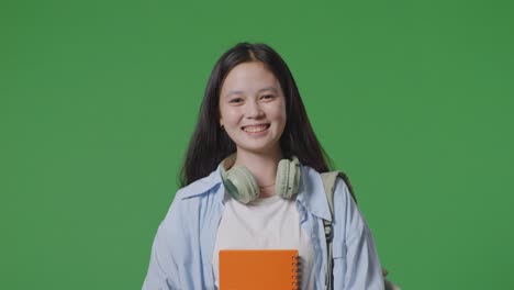close up of asian teen girl student with a backpack holding some books and smiling while walking in the green screen background studio