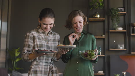 two happy women standing with food in the living room looking and smiling at camera