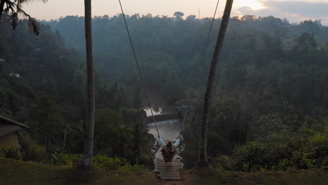 Luftaufnahme-Einer-Frau,-Die-Bei-Sonnenaufgang-über-Dem-Tropischen-Regenwald-Schwingt,-Auf-Einer-Schaukel-Mit-Blick-Auf-Den-Fluss-Sitzt-Und-Spaß-An-Der-Urlaubsreisefreiheit-Hat