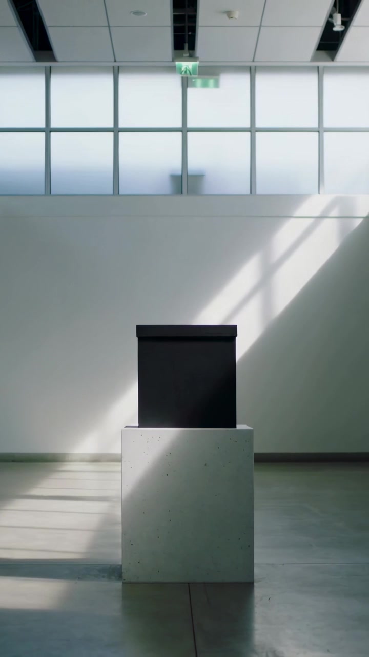 Vertical video: Shifting sunlight sliding across dark box on plinth in gallery, moving soft shadows
