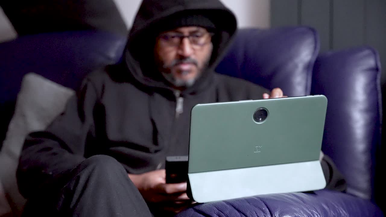 Hooded Asian Man With Glasses Using Tablet Indoors On Couch Holding Phone Technology Communication And Relaxation Concepts