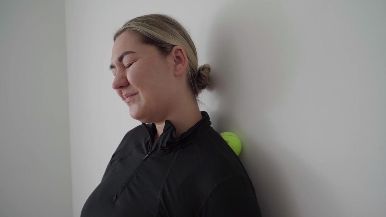 Mulher pressiona uma bola de massagem entre o pescoço, a parte superior das costas e uma parede para aliviar a tensão muscular durante uma sessão tranquila em casa