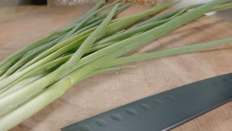 Tiro-Deslizante-De-Cebollas-Verdes-Y-Un-Cuchillo-De-Chef-Negro-En-Una-Tabla-De-Cortar-De-Madera