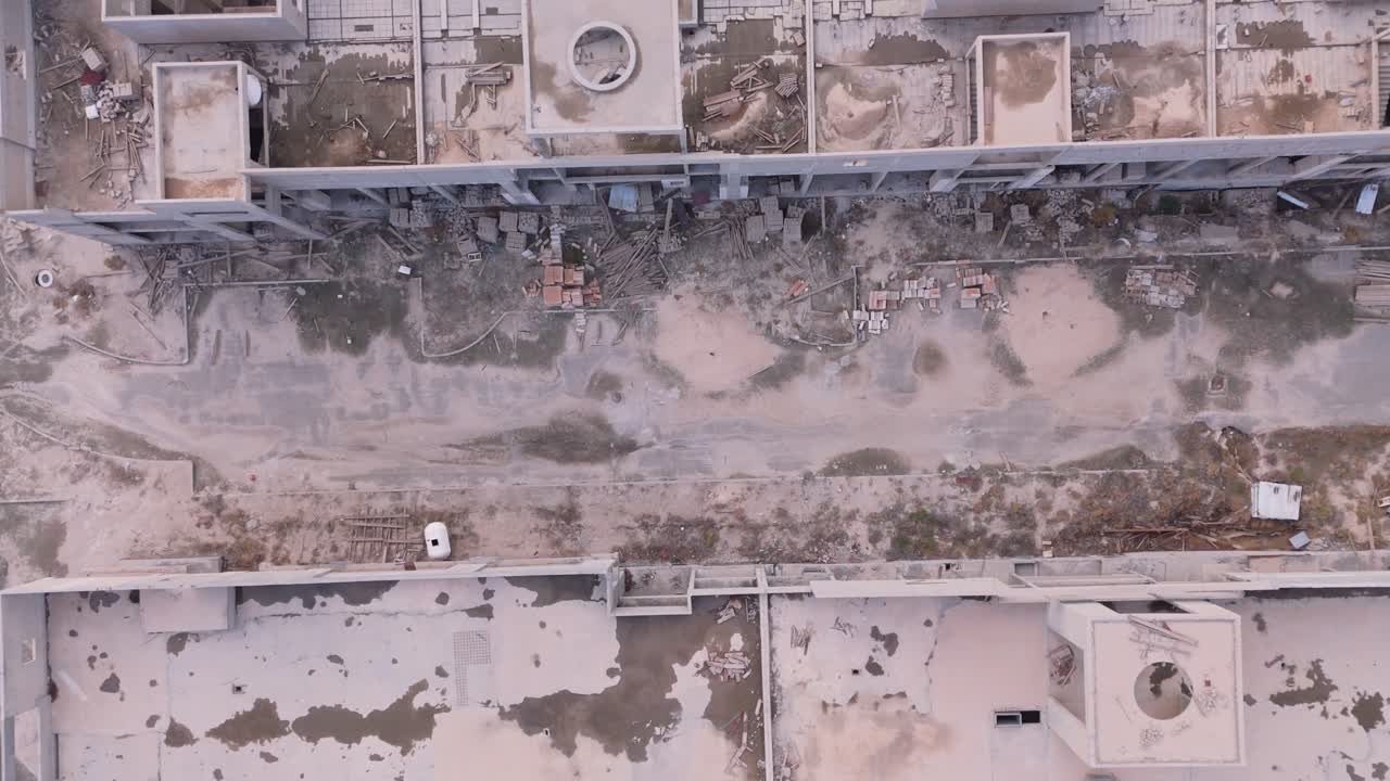 Drone view of a huge unfinished shopping mall or office complex. Features stagnant green water pools, rusted rebar, and overgrown pathways, depicting total economic failure and urban decay