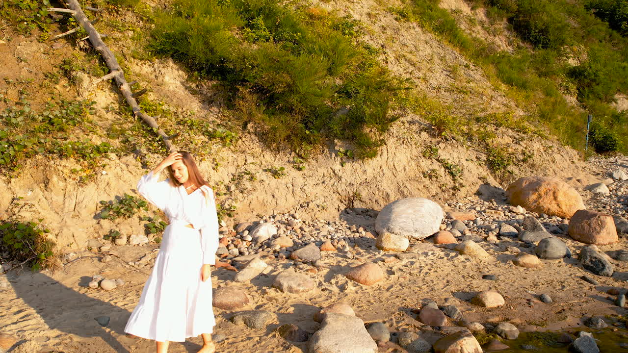 ⁇  드레스를 입은 젊은 여성이 해가 뜨는 동안 몇 개의 큰 바위와 함께 모래 해변의 가장자리를 따라  ⁇ 고 있습니다. 배경에는 바다로 내려가는 절벽이 있습니다.