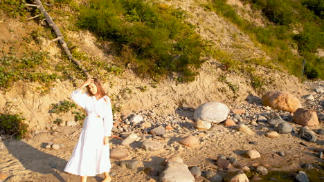 a young woman in a white dress walks along the edge of a sandy beach with a few large rocks during sunrise - in the background, a cliff descending to the sea