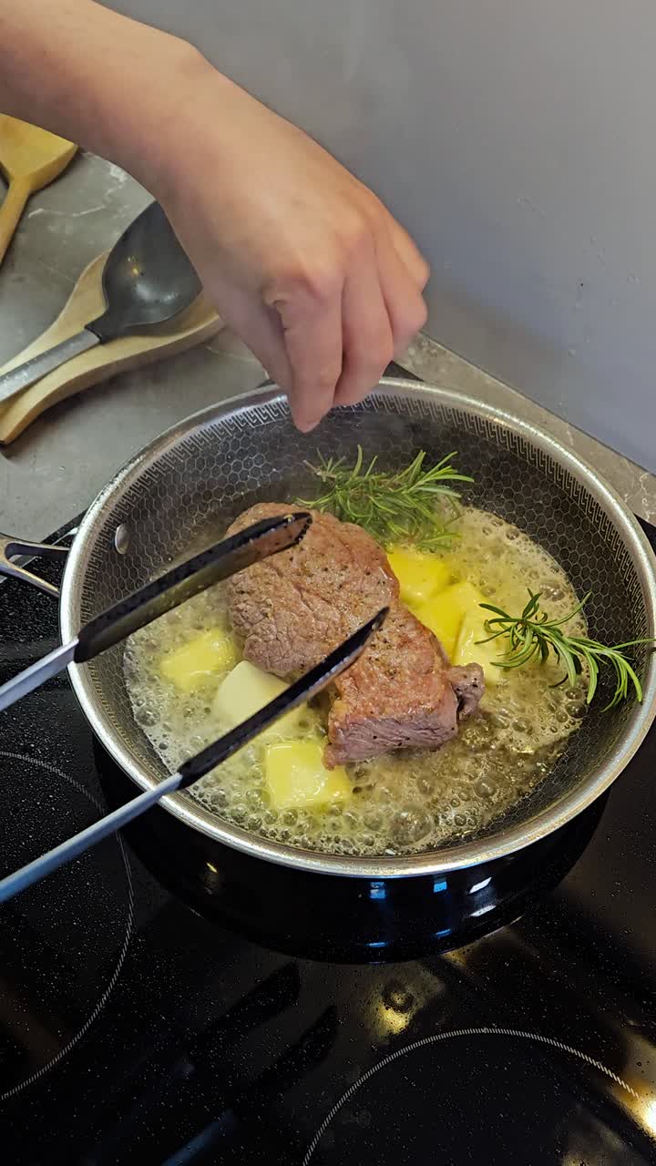 Static vertical shot shows a cook searing a ribeye in hot oil, then dropping butter cubes and fresh rosemary to baste in a stainless skillet; tongs flip the steak as fat sizzles to finish