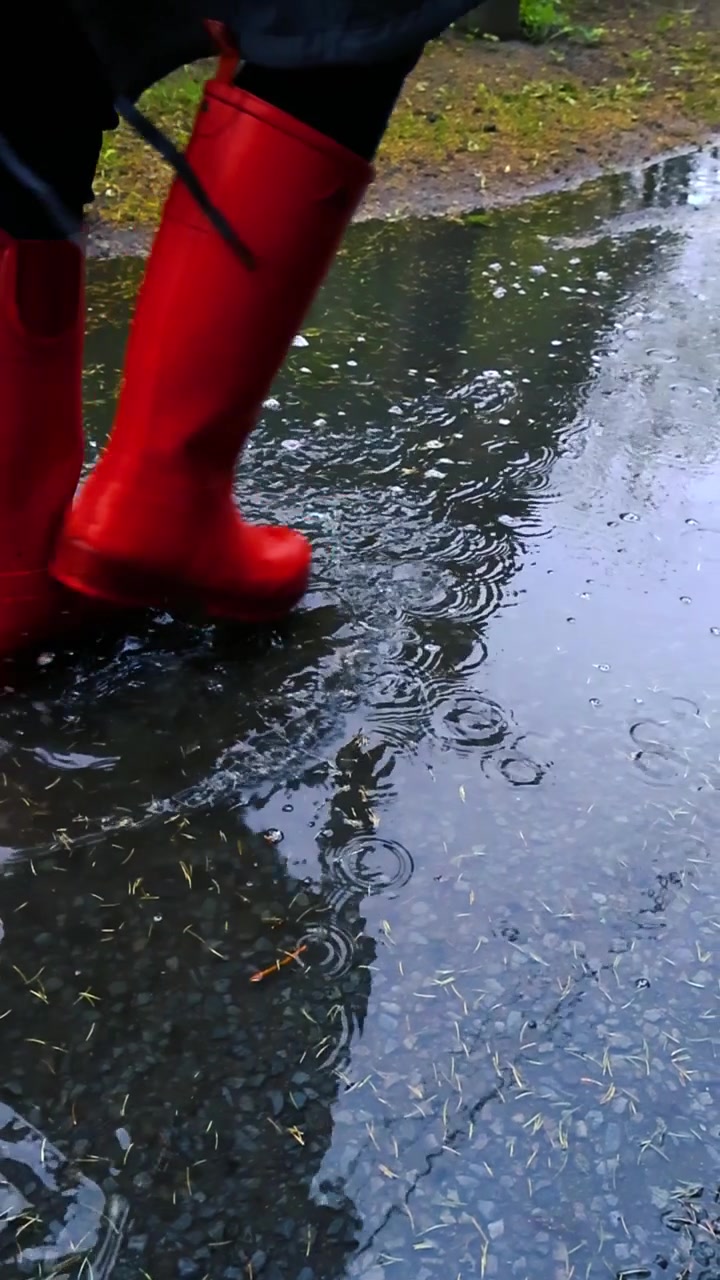 Close up of a person wearing red rain boots walking and splashing through a puddle on a wet asphalt road during a downpour, with raindrops creating ripples on the water surface