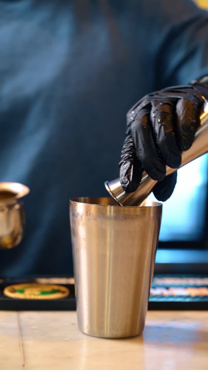 Barman che versa liquido da un jigger di metallo in uno shaker da cocktail. Primo piano delle mani del barman con guanti neri che usa il jigger per misurare gli alcolici nello shaker d'acciaio sul bancone del bar.