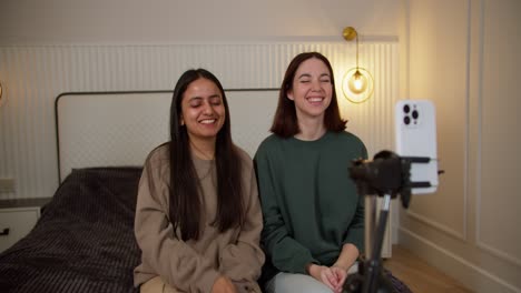 dos chicas morenas felices en un suéter gris y verde están grabando videos de moda para su blog usando un teléfono inteligente blanco mientras están sentadas en la cama en un apartamento moderno en el dormitorio