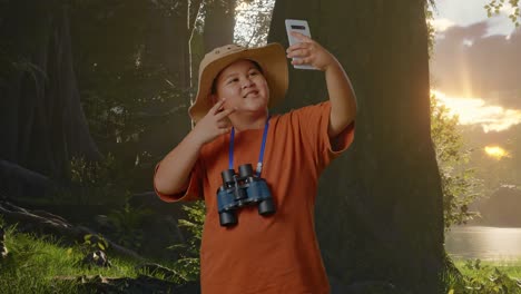 niño asiático con sombrero y binoculares tomando una selfie en su teléfono inteligente mientras explora la naturaleza forestal. niño investigador examina algo, concepto de aventura de turismo de viaje