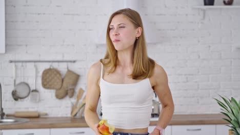 beautiful woman eating tasty apple indoors. young female with sporty body standing in the modern kitchen and eats fresh fruit. portrait of a lovely girl looking aside.