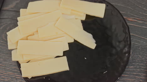 close up of person hand lifting pieces of sliced yellow cheese from black ceramic plate on wooden kitchen counter with visible food crumbs and natural light