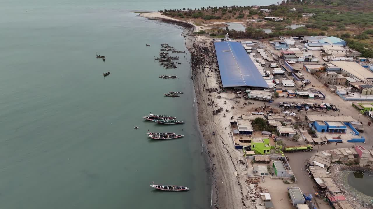 High angle drone view of Joal Fadiouth fishing port, Senegal, with dozens of boats docked by the beach and the lively local market area visible nearby
