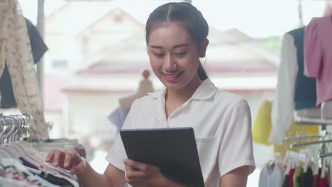close up of asian female owner of clothing store enjoys using digital tablet to check stock on rails