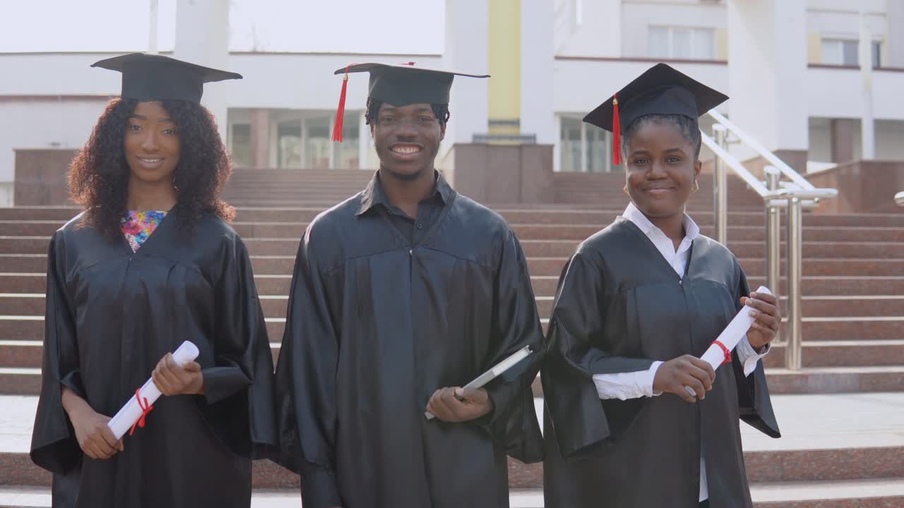 Three African-American university graduates stand outside against the stairs. Students are dressed in the festive uniform of the master